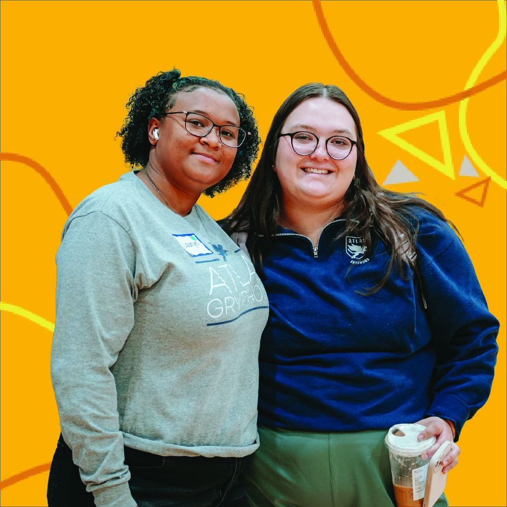 Two PPSC students smiling and standing together in front of a bright yellow background with abstract orange and triangle designs.
